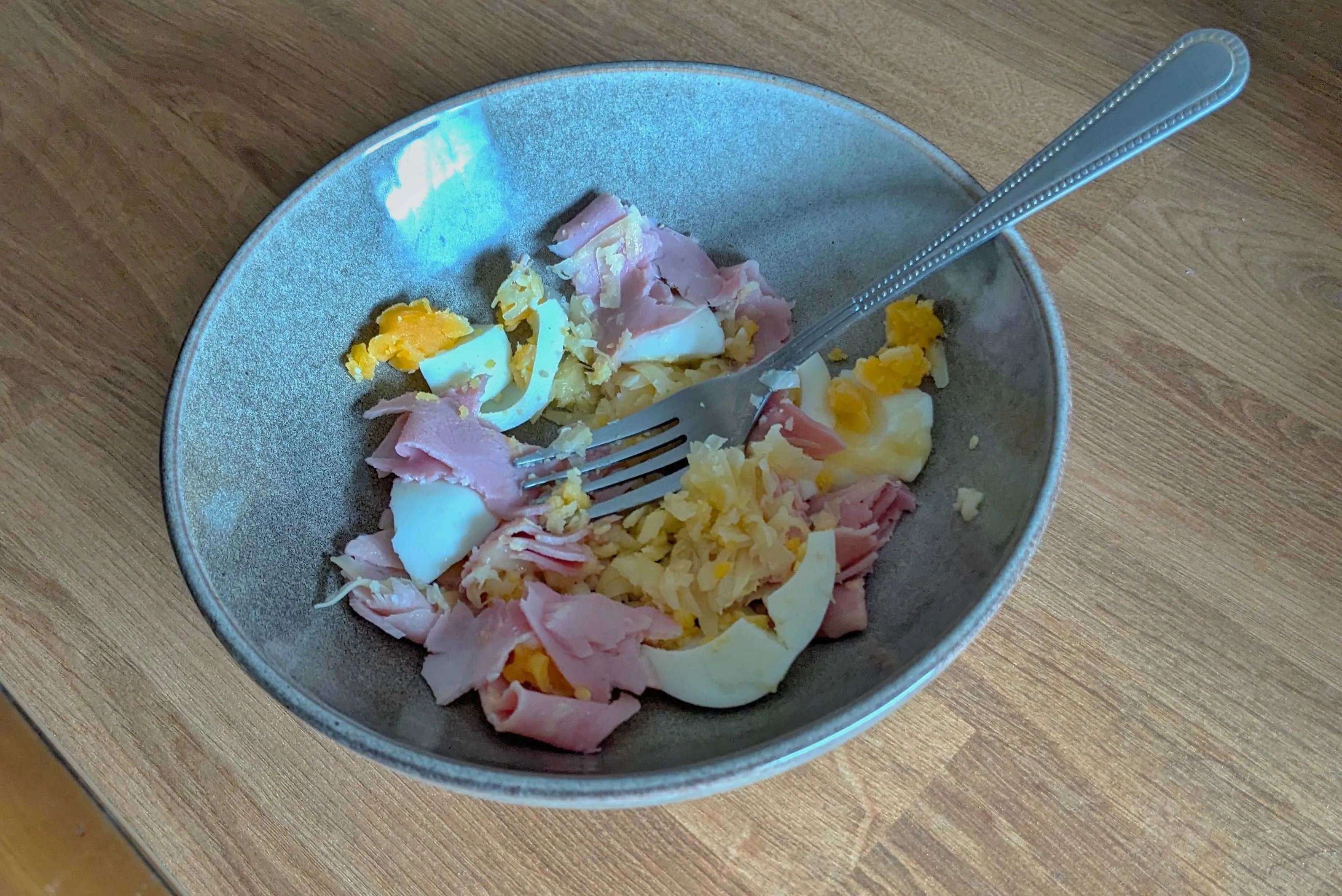 A bowl on a desk. In the bowl is a mash of egg, ham slices, and grated cheese.